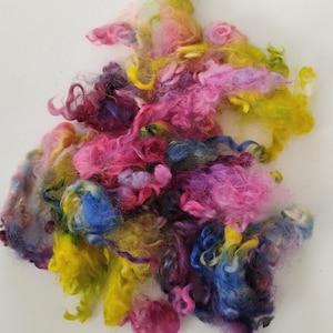 May include: A vibrant pile of colorful fiber in shades of pink, yellow, blue, and purple. The fluffy texture suggests it's likely wool or a similar material, perfect for crafting projects like felting or spinning. The colors are blended.