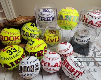 May include: A collection of personalized softball baseballs with embroidered names and numbers. The baseballs are displayed in clear plastic boxes and on a wooden surface. Some of the baseballs are yellow, white, and red.