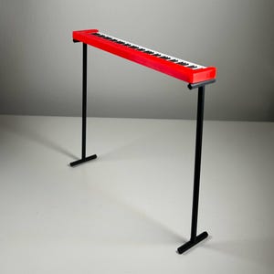 May include: A red toy piano with black and white keys, supported by two black metal stands. The piano's keys are arranged in a standard keyboard layout. The stands have a T-shaped base for stability.