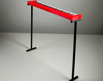 May include: A red toy piano with black and white keys, supported by two black metal stands. The piano's keys are arranged in a standard keyboard layout. The stands have a T-shaped base for stability.