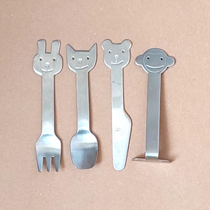 May include: Four stainless steel children's utensils with animal-shaped designs. The set includes a fork with a bunny head, a spoon with a cat head, a knife with a bear head, and a monkey-shaped utensil.