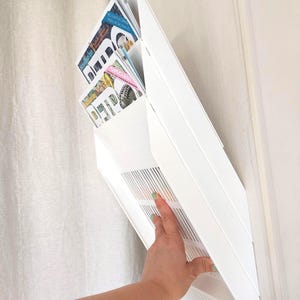 May include: A white wall-mounted magazine rack with two tiered pockets holding several magazines. The rack features a solid white frame with a slatted design on the lower section. The magazines inside display various text and colorful graphic imagery.