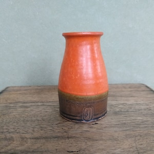 Puede incluir: Un pequeño jarrón de cerámica naranja y marrón con cuerpo redondeado y abertura estrecha. El jarrón tiene una banda marrón alrededor del medio y una base marrón.