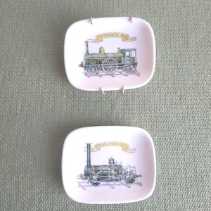 May include: Two small, square, white ceramic plates with rounded corners. Each plate features a detailed illustration of a vintage train. One plate has the text "SPINNROCKEN 1862" and the other has "FORSTLINGEN 1853".