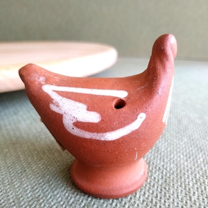 Op de afbeelding: Een kleine vogelvormige fluit van terracotta-kleurige klei met een rond lichaam, een voetstuk en een gestileerd wit geschilderd vleugelontwerp. De fluit heeft een zichtbaar rond luchtgat aan de zijkant en een gladde, matte afwerking.