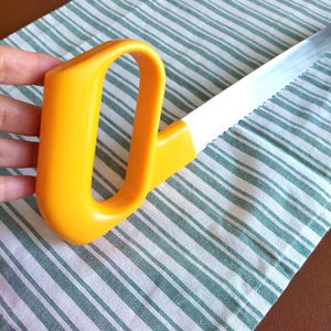 Puede incluir: Una sierra de mano con un mango de plástico amarillo brillante de diseño ergonómico cerrado y una hoja larga, recta y de color plata metálico con un borde dentado. La herramienta está colocada en diagonal, mostrando el contraste entre el vibrante mango amarillo y la hoja de acero reflectante.