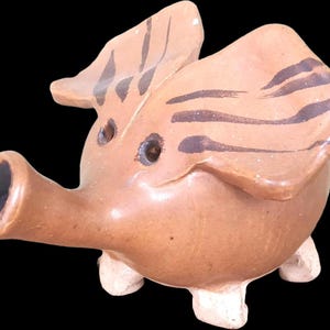 Peut inclure: Figurine de cochon en céramique marron avec des rayures marron foncé sur les oreilles. Le cochon a un long museau et de petits pieds blancs. La figurine est sur fond noir.