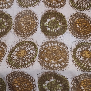 Puede incluir: Una tela blanca con un patrón repetitivo de círculos marrones y verdes. Los círculos están formados por círculos más pequeños y superpuestos que crean un aspecto texturizado.