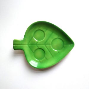 Può includere: Un piatto in ceramica verde brillante a forma di foglia con tre rientranze circolari. Il piatto ha una finitura lucida e un manico a forma di stelo. Il design imita una foglia naturale, ideale per servire snack o come elemento decorativo.