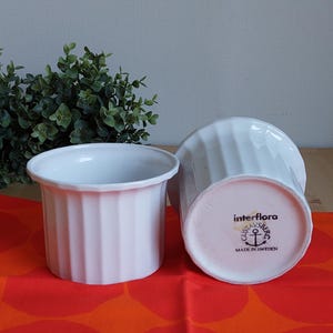 May include: Two white, ribbed ceramic bowls. One bowl is upright, the other is tilted, revealing the "interflora" stamp and "Made in Sweden" text. The bowls sit on a red and orange patterned surface.