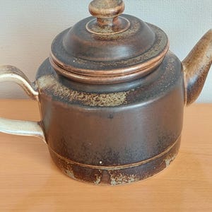 Può includere: Una teiera in ceramica marrone con un coperchio e un manico. La teiera ha una glassa marrone e verde screziata.