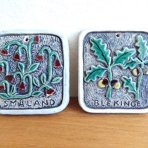 May include: Two square ceramic wall plaques with rounded corners. The left plaque features red bell-shaped flowers on green stems with the text SMALAND. The right plaque displays green oak leaves and brown acorns with the text BLEKINGE.