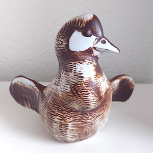 May include: A ceramic bird figurine with a brown and white speckled design. The bird has outstretched wings and a detailed feather pattern. The beak is white, and the eyes are not visible. The figurine is on a white surface.