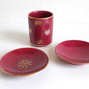 Könnte beinhalten: Eine tiefrote zylindrische Tasse und zwei passende Untertassen, alle mit goldfarbenen Rändern. Die Tasse ist mit kleinen goldenen fächerförmigen Motiven verziert, während eine Untertasse ein zentrales goldenes Sternmuster aufweist und die andere schlicht ist.