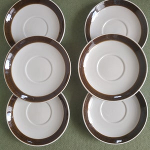 May include: Six brown and white ceramic saucers with a decorative brown band around the edge. The saucers are stacked in two groups of three.