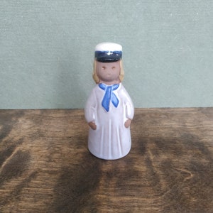 May include: A small ceramic figurine of a girl wearing a white dress, a blue sailor collar, and a blue and white sailor hat.