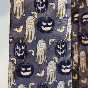 Op de afbeelding: Een stropdas met Halloween-thema met een herhaald patroon van lachende pompoenen, vriendelijke geesten en vleermuizen op een donkergrijze achtergrond. De pompoenen zijn marineblauw met groene stelen, de geesten lichtgrijs.
