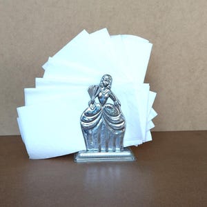 Peut inclure: Un porte-serviettes en métal argenté représentant une silhouette en robe large tenant un éventail. L'objet contient une pile de serviettes en papier blanc unies, disposées en éventail derrière la figurine métallique sur un socle rectangulaire.