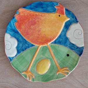 Puede incluir: Un plato de cerámica con un diseño pintado a mano de una gallina amarilla con detalles naranjas de pie en un campo verde con un huevo amarillo. El fondo es azul con nubes blancas.