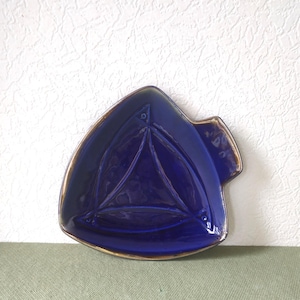 May include: A deep blue ceramic dish with a triangular shape and rounded edges. The surface features a raised, embossed triangular pattern in the centre, and the outer rim is finished with a metallic bronze-toned glaze trim.