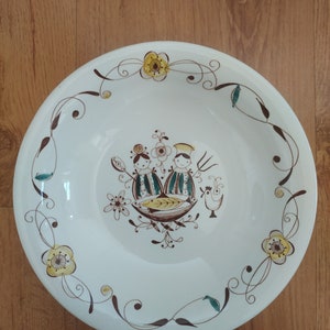 以下が含まれることがあります： 茶色と緑色の花柄が施された白い陶器のボウル。ボウルには、魚と雄鶏を持った2人の人物のスタイリッシュなイラストが描かれています。