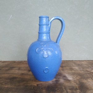 Könnte beinhalten: Ein leuchtend blauer Keramikkrug mit bauchigem Körper und einem einzelnen Henkel. Die Oberfläche weist eine strukturierte Textur mit geprägten Blumen- und Kreismotiven auf. Der Hals ist zylindrisch mit horizontalen Rillen und der Boden ist flach.