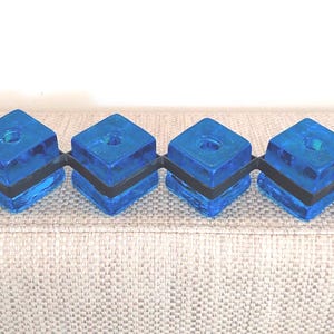 May include: A blue decorative object with four square blocks connected by a black band. Each block has a hole in the centre and is attached to a black loop on each end. The object is on a beige surface.