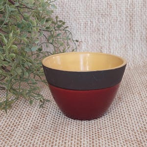 Puede incluir: Un cuenco de cerámica hecho a mano con un exterior rojo texturizado, una banda negra y un interior amarillo. El cuenco tiene una forma pequeña y redonda, ideal para servir aperitivos o salsas. El fondo es una tela tejida neutra.