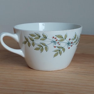 May include: White ceramic teacup with a curved handle. The cup has a green and red floral pattern around the rim. The cup is on a wooden surface.