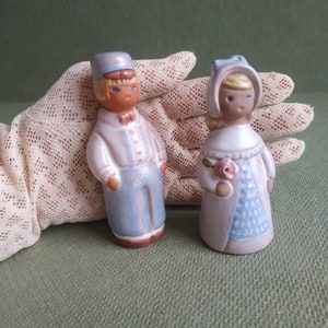 May include: Two ceramic figurines of a boy and girl dressed in blue and white clothing. The boy is wearing a blue hat and the girl is holding a pink flower.