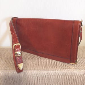 May include: A reddish-brown leather messenger bag with a long adjustable strap and a buckle closure. The bag has a rectangular shape with a flap and gold-colored metal accents on the corners and buckle. The strap is attached to the bag with metal hardware.