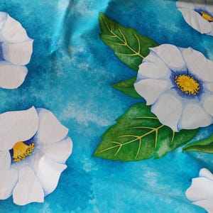 Puede incluir: Flores blancas con centros amarillos sobre un fondo azul con hojas verdes. La tela tiene un efecto acuarela.