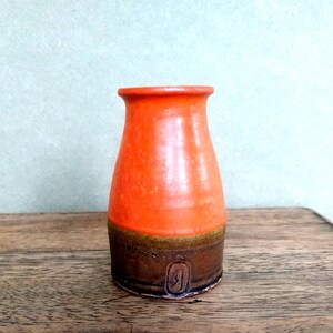Peut inclure: Petit vase en céramique fait à la main, avec une glaçure orange vif sur le dessus et une base marron foncé. Le vase a un sommet légèrement évasé et un corps arrondi. Un petit motif estampé est visible près de la base.