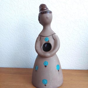 May include: A brown ceramic figurine of a woman with a bun, wearing a dress decorated with blue floral accents. The figurine has a hollowed-out centre and is standing on a wooden surface.