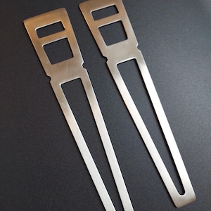 May include: Two silver metal hair accessories with rectangular cutouts. The hair accessories have a long, slender design with a rounded bottom. The accessories are laying on a dark surface.