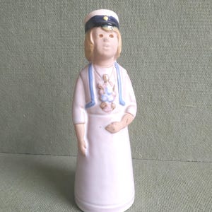May include: A ceramic figurine of a person in a white graduation gown and a white cap with a dark blue band. The figurine has blonde hair and blue gown accents. A small pendant hangs from the neck.