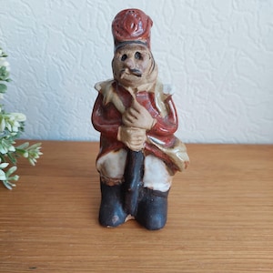 May include: A ceramic figurine of a soldier in a red coat, black boots, and a tall hat. The figurine has a mustache and is holding a rifle. The figure is standing on a wooden surface.