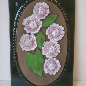May include: A ceramic wall tile with a green border and a brown oval centre. The oval features a cluster of pink and white flowers with green leaves.