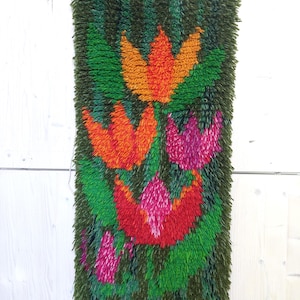May include: A vintage, rectangular wall hanging with a floral design. The rug features a dark green background with vibrant orange, red, and pink flower shapes. The flowers are surrounded by green leaves. The rug is hanging from a wooden rod.