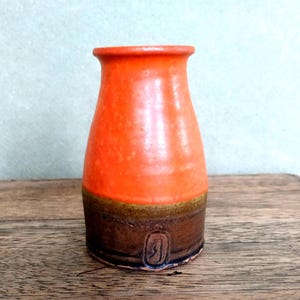 May include: A ceramic vase featuring a bright orange upper section and a dark brown lower section. The vase has a flared rim and a cylindrical body that tapers slightly toward the top. A small, rectangular impressed mark is visible on the base.