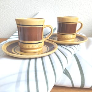 Puede incluir: Dos tazas de café y platillos de cerámica. Las tazas son de color beige con una banda marrón y una raya negra. Los platillos son de color beige con un anillo marrón. El juego está sobre un paño blanco y verde a rayas.