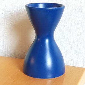 May include: A solid dark blue egg cup with a smooth matte finish and a distinct hourglass shape. The object features a wide circular base that tapers inward toward the center before widening again at the top rim to hold a single egg securely.