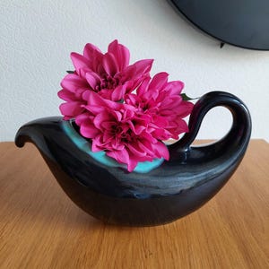 Puede incluir: Un recipiente de cerámica negra brillante con forma de salsera, asa curva y borde interior de color turquesa vivo. El recipiente contiene tres flores de dalia de color magenta vibrante con pétalos superpuestos, creando un contraste llamativo.