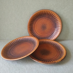 May include: Three brown ceramic plates with a speckled, textured surface. The plates have a dark brown rim and a radial pattern of lines. One plate is oval, and the other two are round.