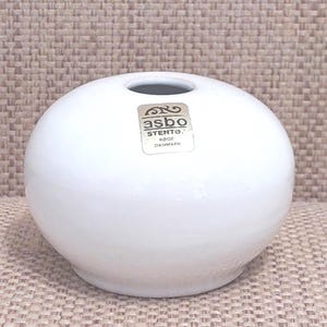 Op de afbeelding: Een kleine, ronde, witte keramische vaas met een enkele opening. De vaas heeft een gladde, glanzende afwerking en een sticker met de tekst "asbo STENTO KOGE DANMARK". De vaas staat op een getextureerd, beige oppervlak.