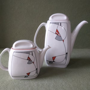 Puede incluir: Dos teteras de cerámica blanca con tapas, con un diseño geométrico en negro, gris y rojo. Las teteras tienen asas y picos curvos. Una es más alta y rectangular, la otra es más corta y cuadrada.