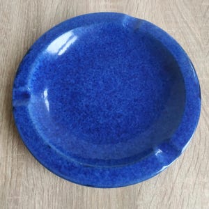 Könnte beinhalten: Ein runder, kobaltblauer Keramik-Aschenbecher mit glänzender, gesprenkelter Oberfläche. Das kreisförmige Objekt verfügt über ein breites, flaches Becken und zwei ausgeprägte Einkerbungen am Rand, die zum Ablegen von Gegenständen dienen. Die Farbe ist tiefblau.