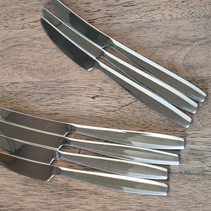 Op de afbeelding: Een set van acht zilverkleurige botermesjes, gerangschikt in twee stapels op een houten oppervlak. De messen hebben een strak, modern ontwerp.