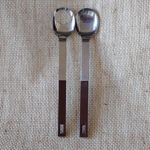 Puede incluir: Dos cucharas de servir de acero inoxidable con mangos marrones. Una cuchara tiene un cuenco ovalado estándar, la otra tiene un recorte circular en el cuenco. Los mangos son rectangulares con un acabado marrón y un pequeño detalle rectangular en el extremo.