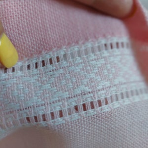 Op de afbeelding: Close-up van een roze gebreide stof met een witte gehaakte rand. De stof heeft een delicaat, getextureerd patroon.